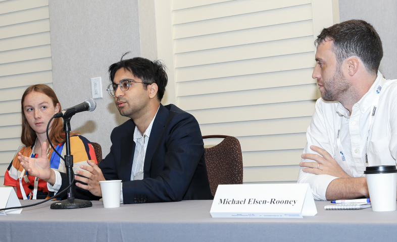 A panel of speakers is seated at a table. One speaks into a microphone.