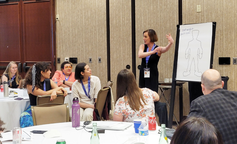 A speaker demonstrates and points at a graphic to a seated group of attendees.